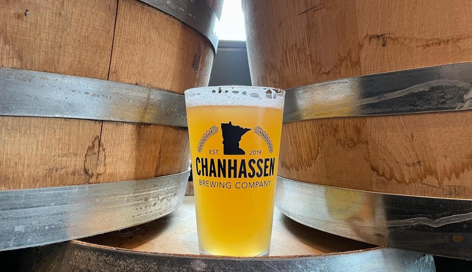 Pint of Beer between two barrels, with the Chanhassen Brewing Co. Logo in the middle